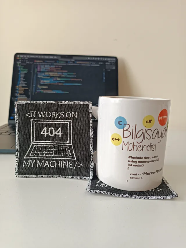 Makine nakışıyla dizüstü bilgisayar ve “It works on my machine” yazısı işlenmiş koyu gri bardak altlığı